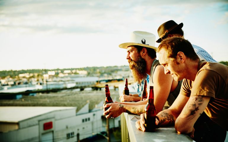 Group of friends leaning on railing on rooftop sharing beers looking out at sunset