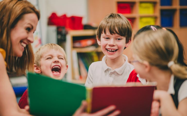 Teacher is sitting in the classroom with her primary school students, reading a story to them.