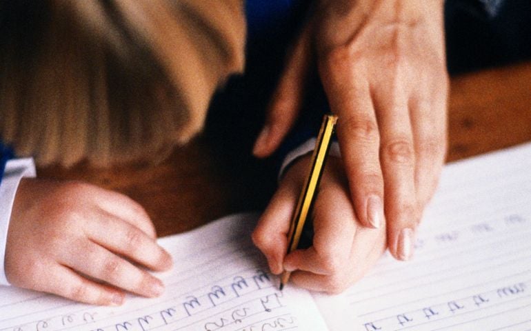 Child (5-7) practising writing in book, guided by adult, close-up