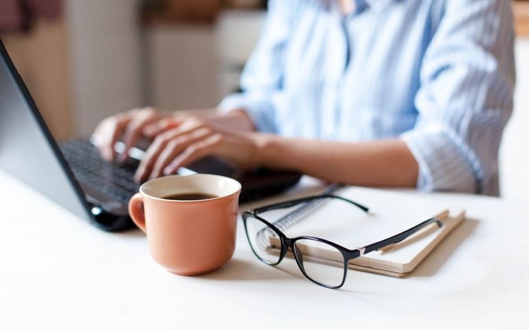 Remote working from home. Freelancer workplace in kitchen with laptop, cup of coffee, spectacles. Concept of distance learning, isolation, female business, shopping online. Close up of woman hands.
