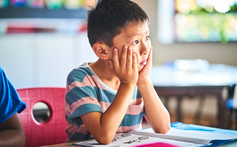 Bored boy looking away at desk. Kid with head in hands sitting in classroom. He is studying in elementary school.
