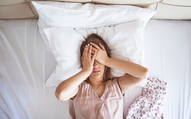 Young beautiful Caucasian woman on bed having headache / insomnia / migraine / stress. Young woman covering her face with hands. Desperate girl suffering insomnia trying to sleep