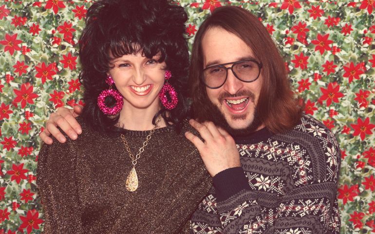 Retro couple portrait at holiday party. 1980s woman smiling, with big curly permed 80s hair wearing large earrings. 1980s man laughing, wearing glasses and a festive holiday sweater. Conceptual image for nostalgia, love, togetherness and relationships.