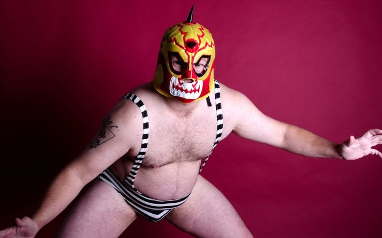 Horizontal studio shot on burgundy of man in Mexican wrestling mask and tiny striped suit.