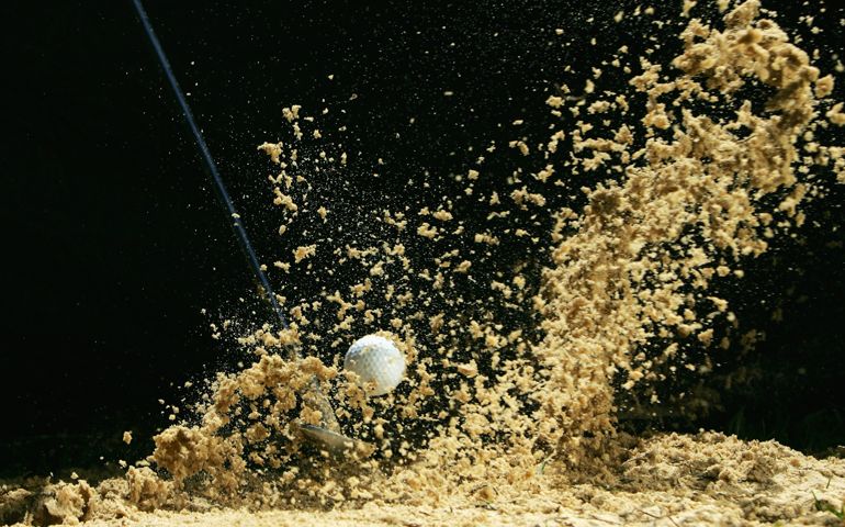 Sand flies up as a golf ball in a bunker is struck by a wedge in front of a black background