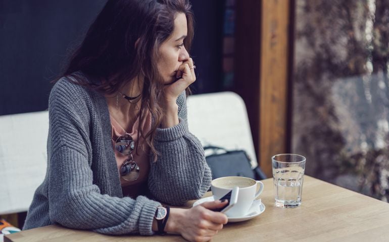Brunette female holding her mobile phone, looking through the window and drinking coffee