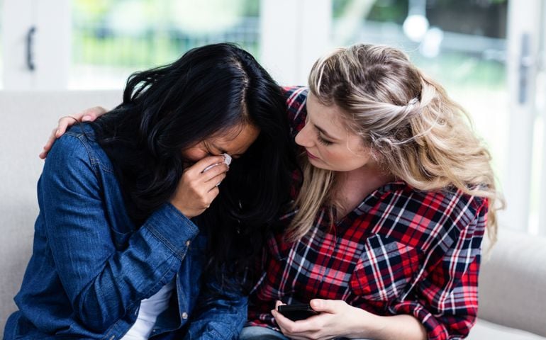 Close-up of young woman consoling crying female friend on sofa at home