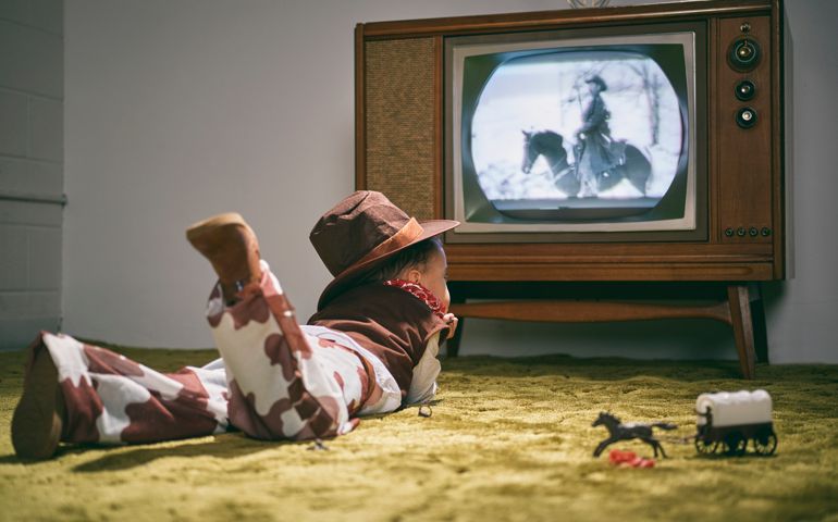 A period correct 1960's television displaying a cowboy scene on the screen (not simulated) with a little boy dressed as a cowboy watching the screen. Image toned to match the era.