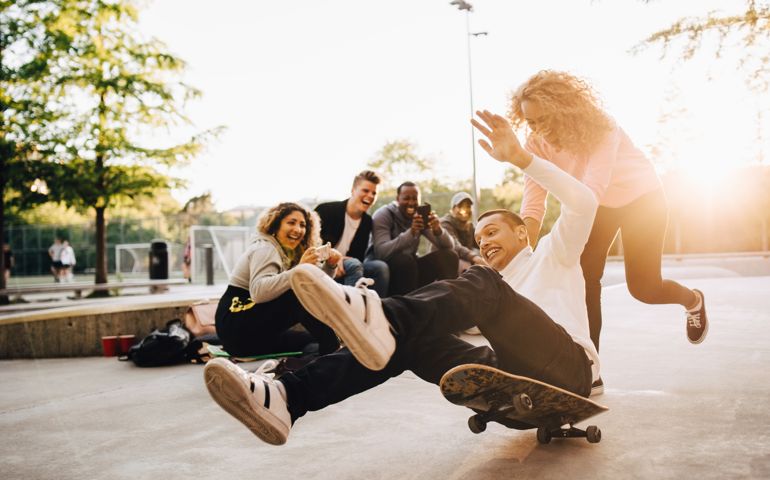 Laughing friends photographing man falling from skateboard while woman pushing him at park