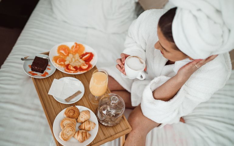 Young woman wearing a bathrobe and a towel, sitting in a bed and drinking coffee and eating healthy breakfast from a wooden tray