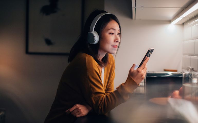 Beautiful and smiling young Asian woman listening to podcast on smartphone at home in the evening. Using smartphone for entertainment.
