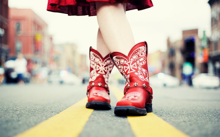 A cowgirl in red cowboy boots and dress has her legs crossed, standing on the double yellow line in the middle of a city street. Ground level view of the boots and legs. Taken in Nashville, Tennessee, USA.