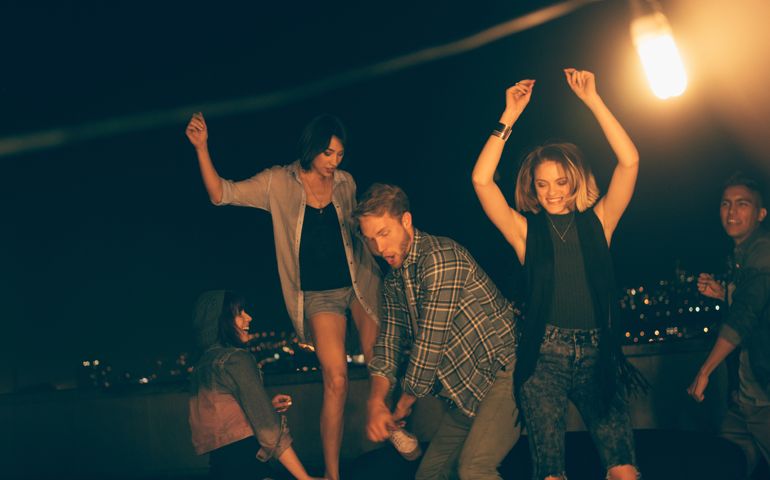 Multi-ethnic group of teenager friends dancing with raised arms and laughing during a night rooftop party