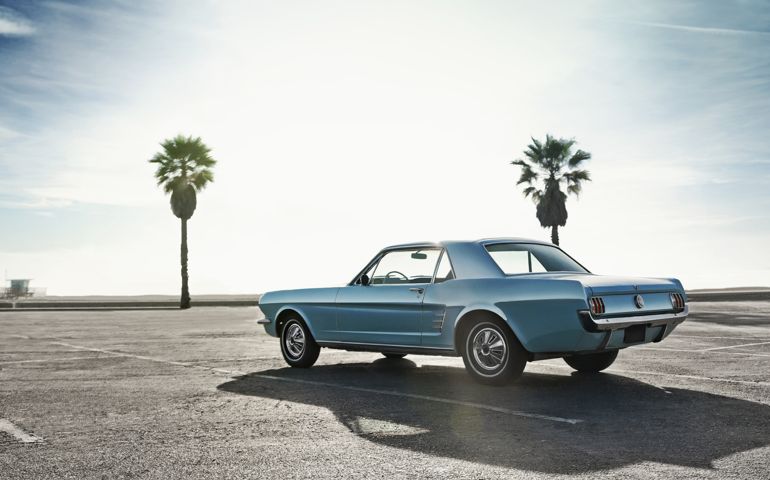Classic blue muscle car on a parking lot close to a beach. Background is: sunny blue sky, two palm trees and sea.