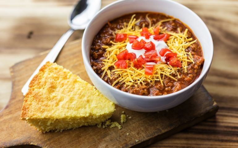 Cornbread and chili, on a wood background with room for copy. Tomatoes, cheese and sour cream toppings are on the chili.