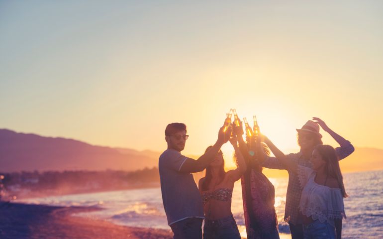 Group of young people partying on the beach at sunset. They are all holding bottles of beer and celebrating with a toast. Silhouette with the beach, sun and sea in the background.