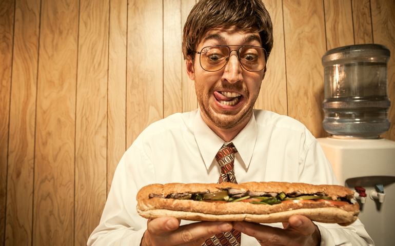 A white collar man working in his office prepares for his lunch; he licks his lips as he looks the large sub sandwich in his hands. Wood paneling in the background, a water cooler visible behind him. Horizontal with copy space.