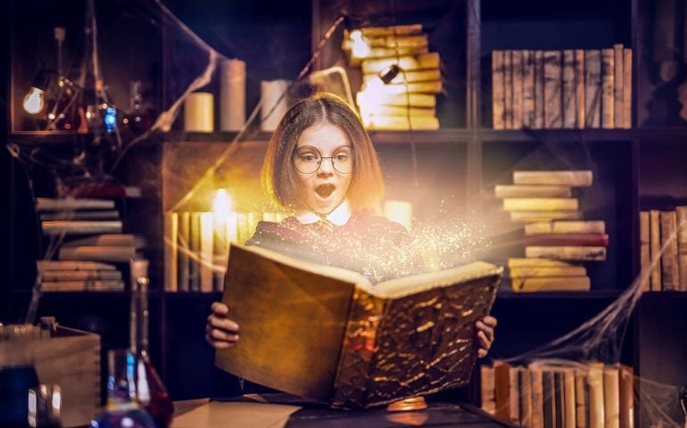 Surprised caucasian little girl in witch Cosplay costume from Harry Potter holds shining magic book