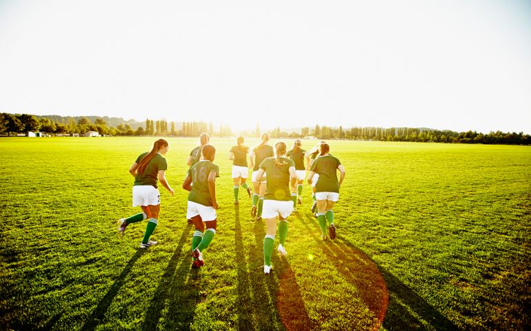 Female soccer teammates running on grass field warming up for soccer match on summer afternoon rear view