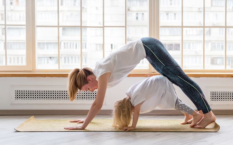 Young mother doing yoga with 3-years girl in front of window. Downward facing dog asana pose