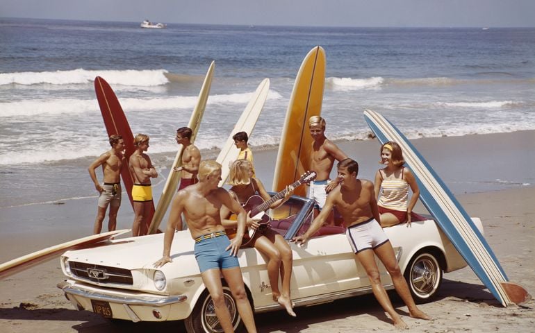 A group of surfers on a beach with a Ford Mustang car.