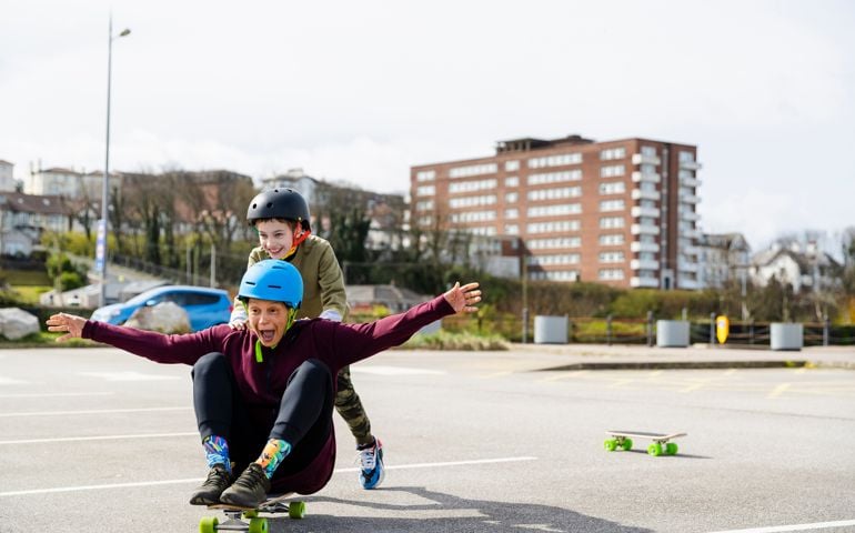 Mid age woman and her son skateboarding in a parking lot in Liverpool in the North of England. The son is pushing his mother who is sitting on the board.