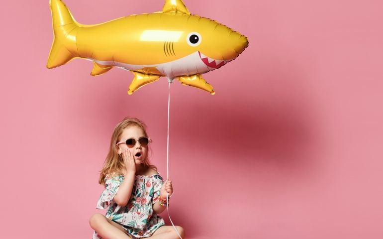 little girl in a summer dress, wears a balloon in the shape of a yellow shark-fis