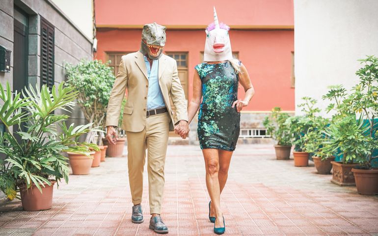 Crazy senior couple wearing dinosaur and unicorn mask - Mature trendy people having fun masked at carnival parade - Absurd, eccentric, surreal, fest and funny masquerade concept