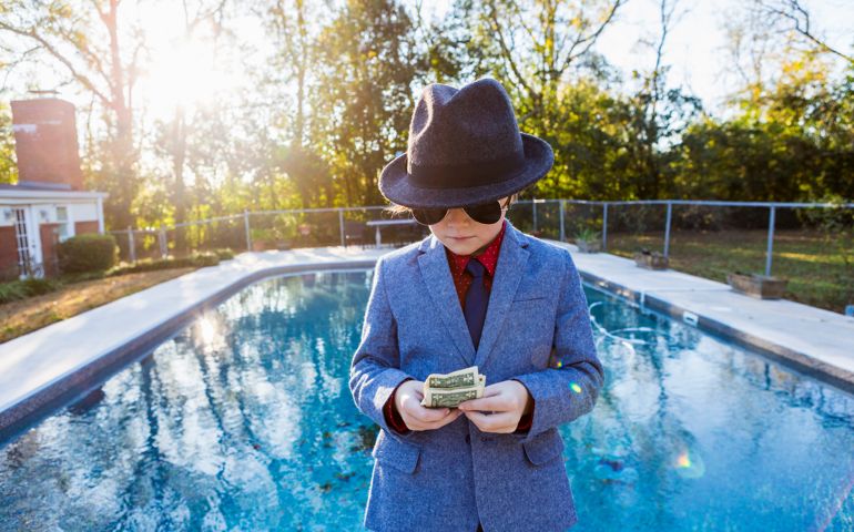 Boy in a suit and dark glasses standing on the edge of water counting dollar bills.