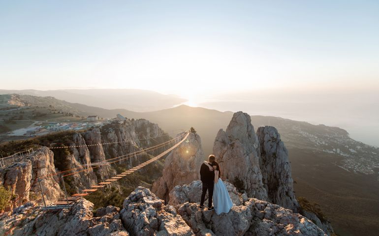 Couple hug each other on the edge of a precipice high above the sea level with a panoramic view of hanging bridge at sunset.
