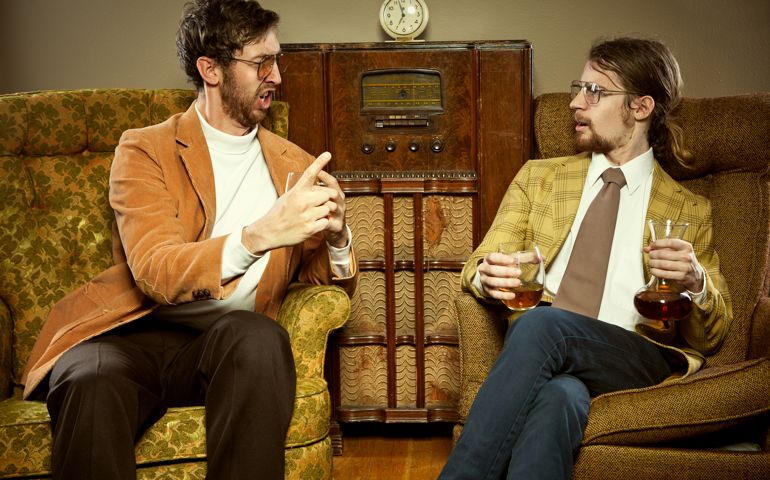 Two men in 1970's or 1980's style clothing sit in a retro living room with cool reclining chairs and an old wooden radio. They both hold glasses of brandy while they gossip, argue, and lecture about important things. Hard wood floors; horizontal.