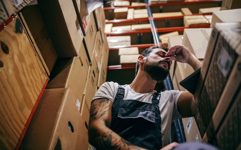 Tattooed bearded worker in overalls having hard day on the work. Storage of import and export firm interior.