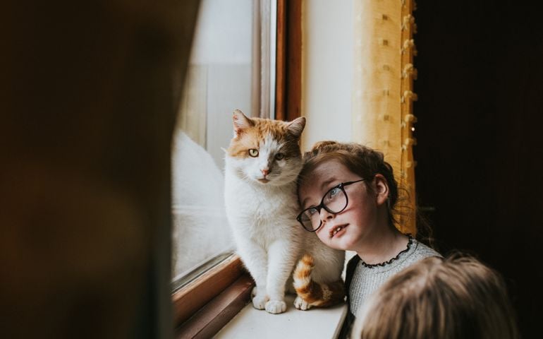 Cute young girl gently rests her head tenderly on her cat. She glances up at him as he sits majestically at a window. The scene is still, quiet, and loving. Curtain provides a space for copy.