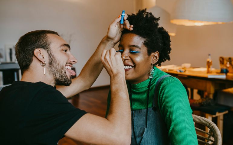 Smiling man applying blue eyeliner to female friend at home