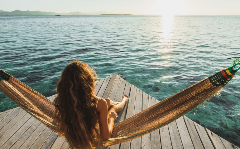 Amazing vacations in Asia. Leisure and relaxation of young beautiful woman in white swimsuit. Hammock on lounge terrace of hotel with over-reef ocean view. Wanderlust travel. Beautiful and natural morning or sunset light.