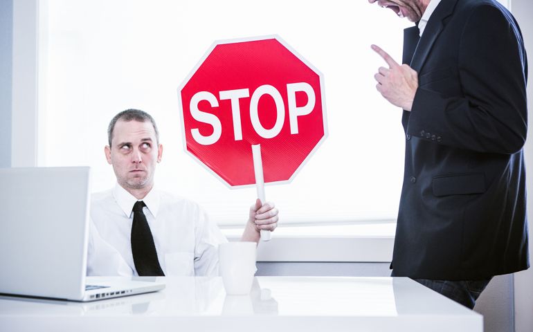 A business man sits at his office desk in front of his computer, while his CEO yells loudly at him. He holds up a stop sign with a funny look on his face, ready to be finished with his job. Horizontal.