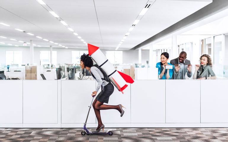Woman speeding thru the office on the scooter with a rocket strapped to her back while her happy multiethnic co-workers are watching her succeed in the workplace. Plenty of copyspace.