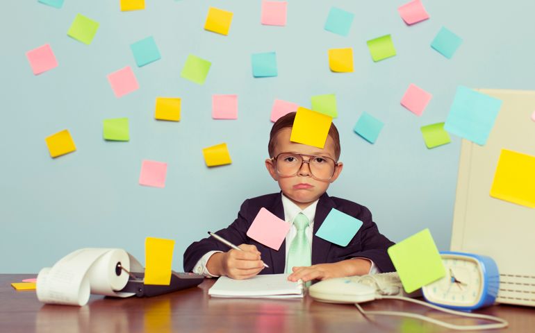 A young businessman sitting at his office computer with pen in hand is completely covered with blank sticky notes. He is forgetting many tasks for his business operations. He wearing a suit and tie and has a frustrated look on his look on his face.