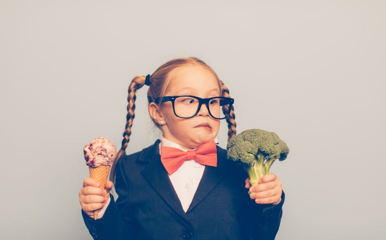 A young female nerd dressed in bow tie and eyeglasses is deciding between eating an ice cream cone or broccoli. She is making a disgusted face at the broccoli. She is choosing the treat.