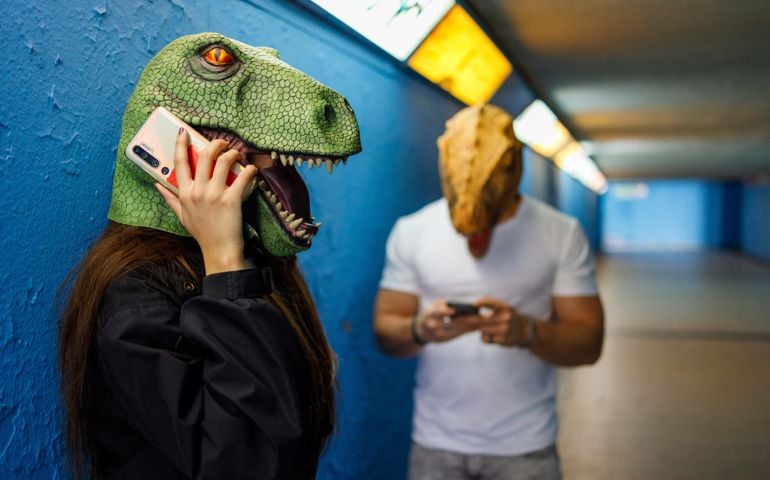 Female talking on smart phone by male friend while wearing dinosaur mask against blue wall