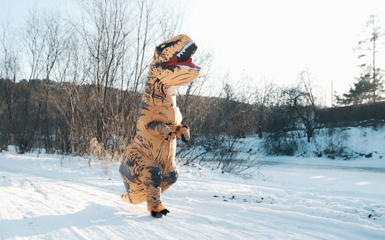 A man in a Tirex dinosaur costume walks through the forest in winter.