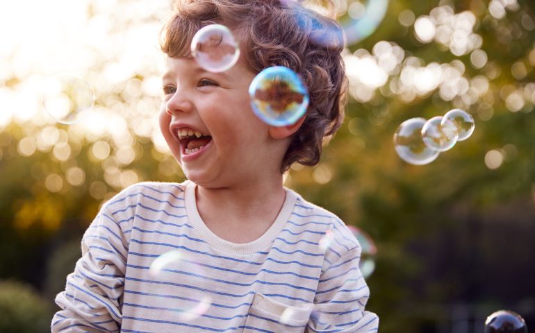 Young Boy Having Fun In Garden Chasing And Bursting Bubbles