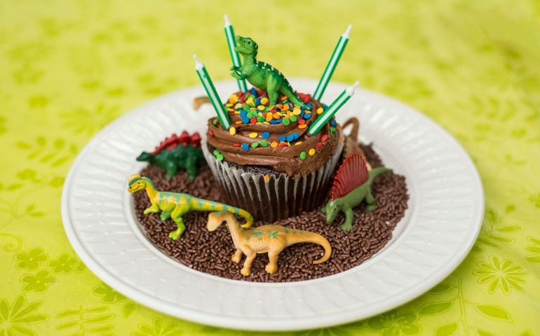 A giant birthday cupcake decorated with plastic dinosaur toys, chocolate sprinkles for dirt, and four candles unlit on the top.