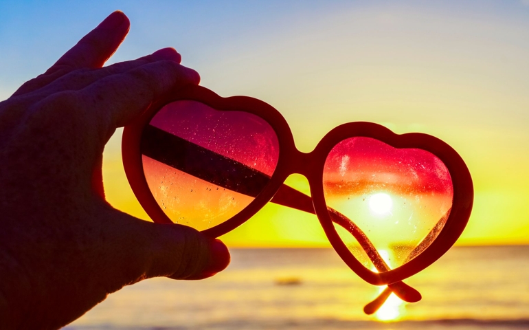Hand holding heart-shaped sunglasses against the sun at beach, summer day