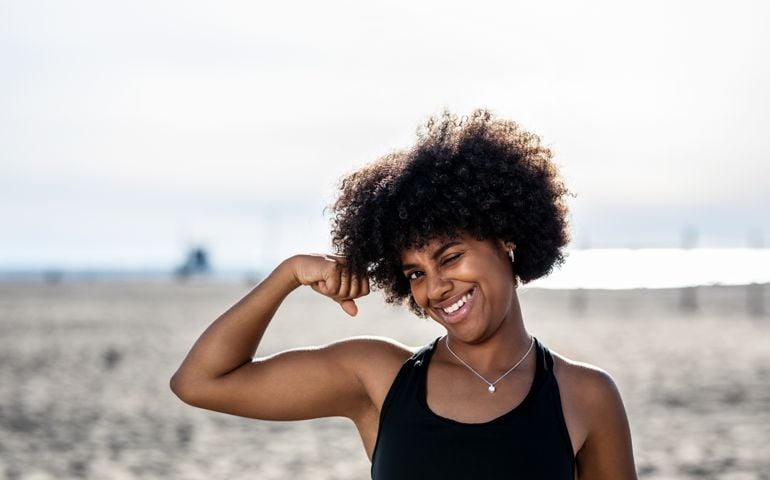 Fit young woman on the beach flexing her biceps. Woman with curly hair wearing sports clothing looking at camera.