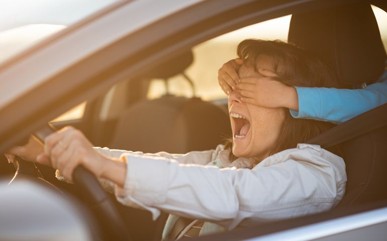 Kid covering eyes of her mother from behind while she is driving car