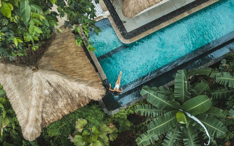 Girl relaxation in private swimming pool hiding in jungle. Wealth travel. Top view, drone point of view directly from above. Vacations in Bali. Tropical and exotic asian nature. Wellness and vitality. Balinese traditional architecture, hotel with straw roof.