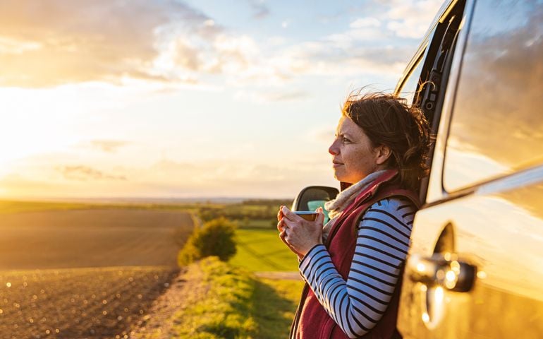 A woman (in her 40s) takes in the view of the countryside from her campervan. She looks contented and relaxed