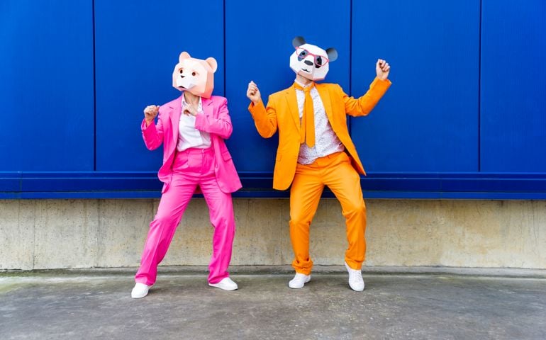 Man and woman wearing vibrant suits and bear masks dancing side by side against blue wall