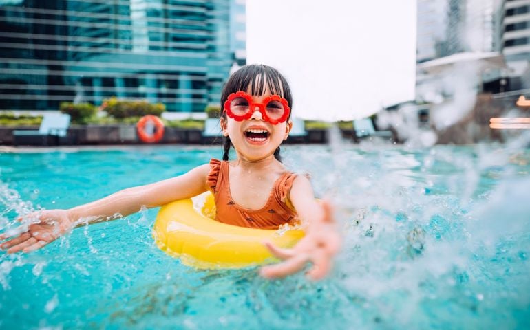 Playful little Asian girl with sunglasses smiling joyfully, splashing and playing with water happily in the swimming pool on Summer vacations